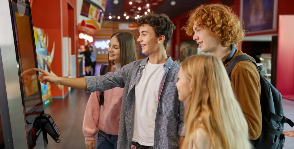 Young friends using self-ordering kiosk at fast food restaurant, smiling while pointing at touchscreen
