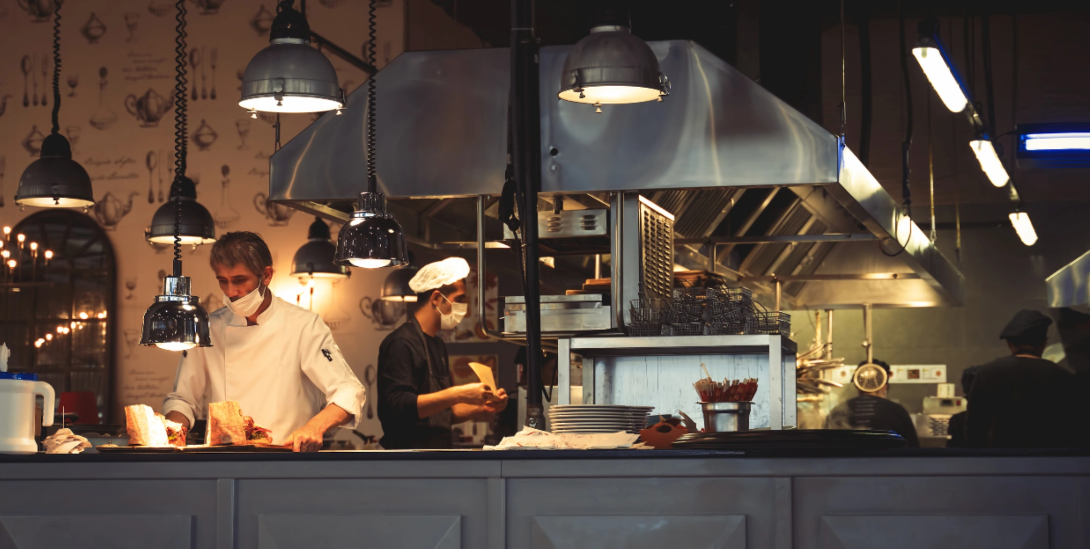 Restaurant open kitchen with pendant lighting, chefs at work behind counter during dinner service
